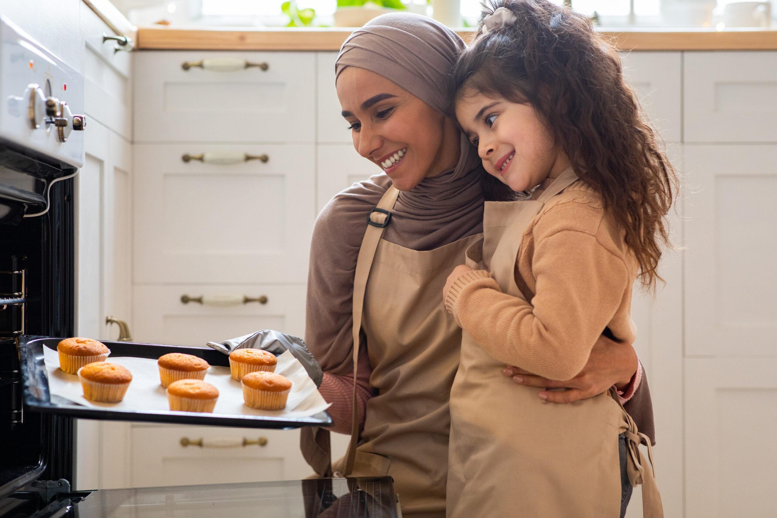 Happy muslim mother and daughter baking in kitchen, holding tray with muffins